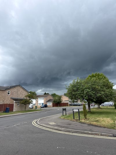 This image captures a residential street scene in Hazlemere, United Kingdom, under a dramatically dark and brooding sky, indicating an approaching storm. The setting is a suburban road lined with brick houses and green spaces.

The foreground features an asphalt road with distinct double yellow lines along the curb, indicating parking restrictions. A grey paved sidewalk runs parallel to the road, separated by a strip of green and somewhat dry grass. Further down the street, the road curves gently to the right. On the left side of the road, a large, leafy green tree stands prominently, its foliage vibrant against the dark backdrop of the clouds. Beyond the tree, a series of brick-built houses with tiled roofs are visible, some featuring white window frames. A dark blue car is parked in a driveway of one house, and a grey refuse bin is visible near another.

On the right side of the road, another row of houses can be seen, partially obscured by the trees and general perspective. A silver van is parked on a grass verge further along the street. A black street sign with white lettering clearly reads "FIRS CLOSE," providing a specific location cue within Hazlemere.

The most striking element is the sky, which transitions from a lighter grey on the far left to a dense, very dark grey mass of heavy clouds across the majority of the frame, suggestive of an imminent thunderstorm or significant rainfall. The overall lighting is subdued, typical of a heavily overcast day. No people are visible in the scene. The image conveys a sense of anticipation due to the dramatic weather conditions.