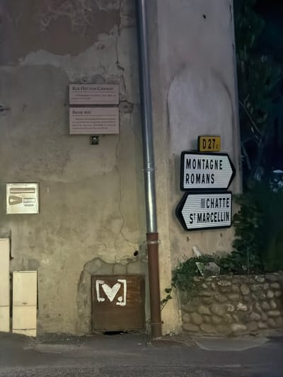 The image shows a section of a textured, aged wall in Saint Antoine l'Abbaye, France. Mounted on the wall are two rectangular plaques with French text, one above the other, followed by a small square emblem. The top plaque reads "RUE HECTOR GARAUD" and the bottom plaque reads "BASSE RUE." Below these, a metal downspout runs vertically along the wall. To the right of the downspout, two directional road signs are mounted. The top sign indicates "MONTAGNE ROMANS" with an arrow pointing right. The lower sign indicates "CHATTE ST MARCELLIN" with an arrow pointing right. Above these signs is a smaller yellow and black road sign with "D 27c". To the left of the downspout, there is a small, square metal object mounted on the wall, possibly a plaque or a utility cover. Further to the left, partially visible, are two light-colored rectangular panels, likely utility boxes or covers. At the bottom of the wall, to the left of the downspout, is a square, rust-colored metal panel with a white graphic resembling a heart shape within a stylized "M". The ground in front of the wall is asphalt. A low stone wall runs along the right side of the scene, with foliage growing over it. The scene appears to be outdoors, at night or in low light conditions, with artificial lighting illuminating the wall and signs.
