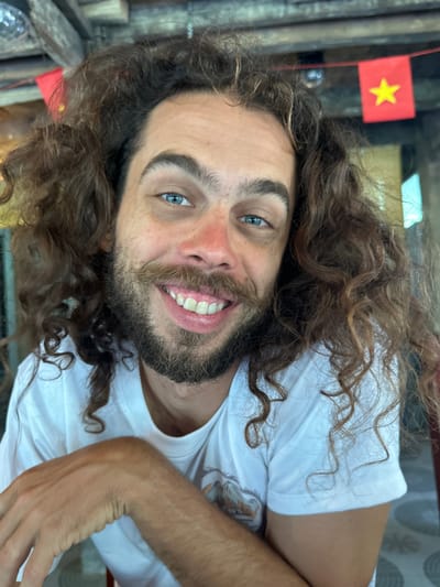The image features a man with long, curly brown hair, striking blue eyes, and a full beard and mustache, captured in a rotated orientation. He is smiling broadly, revealing his teeth, and appears cheerful and friendly, with a slight flush on his cheeks and nose. He is wearing a white short-sleeved t-shirt, parts of which are visible across his torso and arm.

In the upper left background, a small Vietnamese flag is clearly visible. It is red with a yellow five-pointed star, affixed to a thin pole or string, confirming the location as Vietnam, specifically within Du Già Commune as per the context. The background elements behind the man are dark and somewhat indistinct, appearing to be rustic wooden structures or an indoor setting, possibly a local establishment. The lighting suggests it is daytime, with some ambient light illuminating the man's face, while the background remains in relative shadow. There is no discernible text other than the emblem on the flag. The image captures a casual moment, highlighting the man's bright expression against the backdrop of the Vietnamese flag.