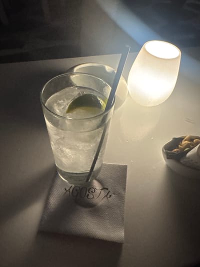 A tall glass filled with ice and a clear liquid, garnished with a slice of lime, sits on a white table next to a small bowl of nuts and a small glowing lamp. A black straw extends from the glass. Underneath the glass is a folded white napkin with embroidered lettering that appears to read "AGOSTA". The scene is dimly lit by the lamp, casting shadows across the table. The background is dark and out of focus, with hints of textured surfaces. The setting is likely an establishment in Naousa, Greece, given the napkin's branding.