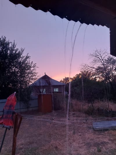 This is an image of a rural scene, likely a dacha or country property in Dachi Su, Kyrgyzstan. The perspective is from under a dark, corrugated overhang, possibly a roof or porch. Several thin streams of water are falling from the edge of this overhang, suggesting it might be raining, or perhaps sprinklers are in use.
The background depicts a twilight sky with hues of soft purple and pink, indicating either dawn or dusk. Silhouetted against the sky are the bare branches of a tree on the left and the dense foliage of trees and bushes on the right. In the mid-ground, a rustic building is visible. It has a dark roof and what appears to be a reddish-brown section, possibly an extension or shed. Windows on the building are dimly lit. A fence runs along the property line.
The ground is covered in dry, straw-like grass. In the foreground, on the right side, a partially unfurled red and black patterned umbrella or tarp is propped up. The overall mood of the image is tranquil and slightly melancholic, typical of the quiet moments at the end or beginning of the day in a rural setting. There is no visible text in the image.