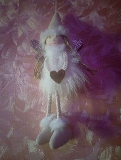 A decorative plush angel ornament hangs against a textured, pink and purple background. The angel has a light-colored face with painted features, a white fuzzy body, and glittery purple wings. It is holding a small brown heart-shaped object. The background appears to be wallpaper or fabric with a subtle, directional pattern. The image is illuminated by light that casts shadows and highlights on the textured surface. The angel ornament is suspended from a string, suggesting it is meant to be hung as a decoration. There are no people or animals present in the image. The scene is indoors, with the lighting suggesting it is not direct sunlight. The location is specified as Меткомелино, Russia.