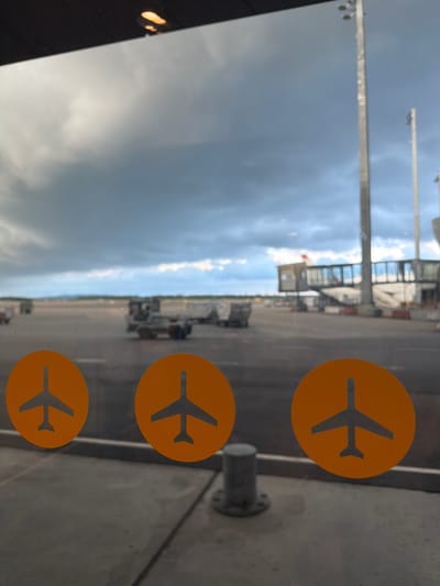 This is a photograph taken through a glass pane, likely at an airport terminal. Three orange circular decals with a black airplane silhouette are visible on the glass in the foreground, arranged horizontally. The decals are partially obscured by reflections.

Beyond the glass, an airport tarmac is visible under a cloudy sky. Several airport vehicles, including baggage carts and a tug, are scattered across the tarmac. In the background, a jet bridge is connected to a building, and another jet bridge extends from a different structure. Tall light poles are also present on the tarmac. The overall scene suggests a view of an active airport environment.

The specific location in Gardermoen, Norway, refers to Oslo Airport, Gardermoen (OSL), which is the primary international airport serving Oslo and the surrounding region.