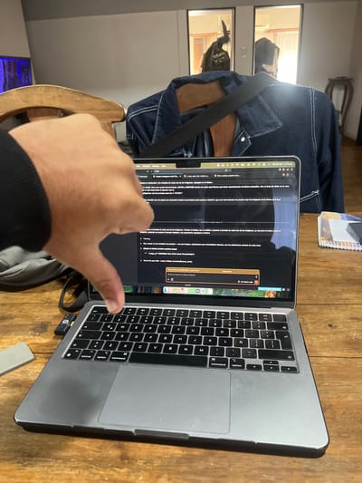 An indoor scene captured from a slightly elevated, side angle shows a person's right hand in the foreground, extended towards an open MacBook Pro laptop. The laptop rests on a light brown wooden desk. A black USB receiver is plugged into the laptop's right side.

The main subject is the laptop screen, displaying what appears to be a conversation or project management interface, likely in an application like Notion, involving AI tools such as Claude and Gemini. The content on the screen is predominantly in Spanish. Visible text includes discussions about image resolutions, measurements, creating prompts for AI image generation ("Gemini y nano-banana"), and ensuring consistent framing for "ANTES Y DESPUÉS" (BEFORE and AFTER) cards. There are references to a "Plan coding session" and "Claude AI Notion". A notification reads, "You've hit your limit resets 2:50pm (America/Buenos Aires)," indicating the time zone and suggesting the user is in Sarandí, Argentina. The top bar of the laptop shows "Tue 28 Apr 11:12am."

In the background, a dark denim jacket with white stitching is draped over a wooden chair. Reflections of windows, possibly indicating an outdoor view or another room, are visible behind the jacket. A small spiral-bound notebook is also on the desk to the left of the laptop. The overall setting suggests a workspace or home office.
