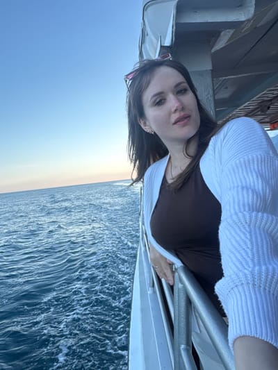 A woman is seen standing on what appears to be a boat or a ferry. She is wearing a brown top and a white cardigan, and she is holding onto a silver railing. Beyond the railing is a view of the sea. The sky is visible above the sea, showing a gradient of light blue and white. The sea itself is a dark blue with white waves.
The location appears to be Гаспринский поселковый совет, Ukraine, given the presence of water and coastal features. The woman is the primary subject, and the sea and sky form the backdrop.