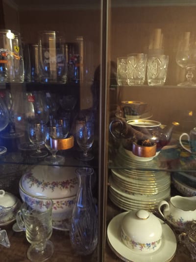 The image shows a glass-fronted display cabinet filled with various tableware and glassware. On the top shelf, to the left, are several clear glasses, one with a red and yellow racing car graphic and a "Ferrari" logo. To the right of these are more clear glasses, some with etched patterns. The middle shelf on the left contains a mix of clear glasses of different shapes and sizes, some with gold rims. On the middle shelf to the right, a dark blue and gold teapot sits on a saucer. Below the teapot are stacks of white plates with gold rims. The bottom shelf on the left features a large, ornate serving bowl with a floral pattern, a smaller lidded dish, a clear glass mug with a handle, and a tall, clear glass vase with a fluted design. The bottom shelf on the right shows a white creamer with a floral pattern and another ornate silver-colored object.
The image does not contain any information that would indicate its location in the city of Pavlohrad, Ukraine.