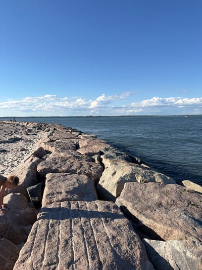 The image depicts a sunny day at a beach in Hampton Bays, New York. A person, a young boy, is seen on the left side of the frame, bending down on a sandy area next to a large stone jetty. The jetty, composed of massive pink and grey granite rocks, extends into the dark blue ocean. The water is calm with small ripples. In the distance, a thin strip of land with trees and some structures is visible, bordering the horizon. The sky above is a clear, bright blue with scattered white cumulus clouds. The sandy beach is visible to the left of the jetty, with footprints imprinted on its surface.