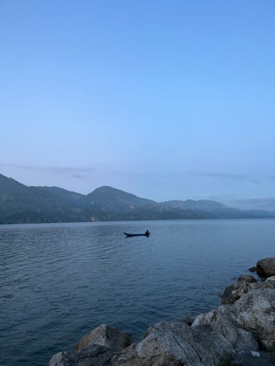In Merodot, Indonesia, a serene lake scene unfolds as the sky transitions to dusk. The water's surface, a deep blue, is gently rippled by a subtle breeze. In the distance, a silhouette of mountains covered in lush vegetation forms a hazy backdrop against the pale blue sky, hinting at the surrounding natural beauty.
Dominating the foreground, a collection of large, weathered rocks sits along the edge of the lake, their rough textures and varied shades of gray and white contrasting with the smooth water. Among these rocks, a splash of plastic debris is visible, a small testament to human presence.
On the lake, a lone blue boat drifts serenely. A single figure, barely discernible in the dim light, sits in the boat, their posture suggesting a moment of quiet contemplation or perhaps an evening fishing endeavor. The boat itself is small and simple, a traditional vessel navigating the tranquil waters. The overall atmosphere is one of peaceful solitude and natural splendor.
