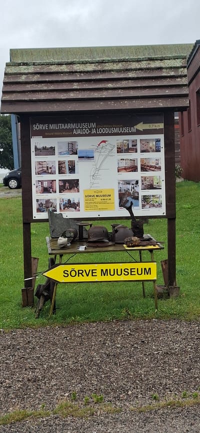 This image shows the Sõrve Military Museum in Sääre, Estonia. The museum is housed in a wooden building with a weathered roof. In front of the building is a sign with information about the museum, including its name, address, and opening hours. The sign also features several photographs of the museum's exhibits. Below the sign, a yellow arrow pointing to the left indicates the direction to the museum, with "SÕRVE MUUSEUM" written on it. A table in front of the arrow displays a collection of historical artifacts, including helmets and what appear to be parts of military equipment. The area around the museum is grassy, with a gravel path in the foreground. A car is parked to the left of the sign. The sky is overcast.