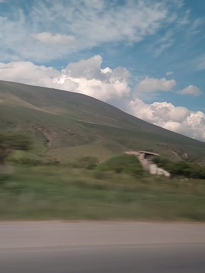 This media file captures a wide landscape view, likely taken from a moving vehicle, given the noticeable motion blur in the foreground and midground. The image is oriented vertically (or rotated 90 degrees), with the sky originally at the top and the ground below.
The upper portion of the image features a bright blue sky with numerous white clouds. Some clouds are large and fluffy cumulus formations, while others are thinner and more scattered, suggesting good weather.
The central and left parts of the image are dominated by a large, undulating green hill or mountain, covered in vegetation. Its slopes rise towards the horizon. In the midground, on a grassy slope to the right, there is a blurry, light-colored structure that appears to be a small building, ruin, or a man-made monument, indistinct due to motion blur.
On the far right, a pale, light-colored road or paved path runs parallel to the edge of the frame, separating the grassy area from the rightmost edge of the image. The foreground grassy area is also blurred, reinforcing the sense of movement.
The scene appears to be set during the daytime, likely late morning or early afternoon, under clear skies with scattered clouds. No people or visible text are discernible. The landscape, with its green hills and road, is consistent with an outdoor scene in a rural or mountainous region like Zangakatun, Armenia.