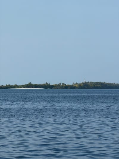 The image depicts a scene in Dhigurah, Maldives, featuring a tranquil body of water. The water's surface exhibits gentle ripples and reflections. A thin strip of vegetation lines the horizon, separating the water from a clear, light-blue sky that dominates the left side of the frame. No people or distinct objects are visible. The overall environment conveys calmness and natural beauty.
