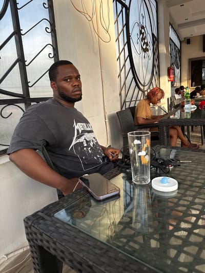 A man is seated at an outdoor glass table. A woman with red dreadlocks is seated at another table behind him, looking at a mobile device. Other people are seated in the background. Keys, a drinking glass with cartoon animal decals, and a small white container with a blue figurine are on the glass table. The location appears to be an outdoor patio or veranda area, with a roof structure supported by iron lattice. The scene is in Rubuchi, Nigeria.