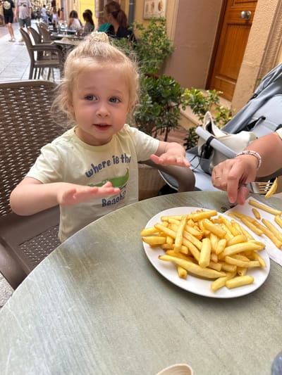 In Sant Feliu De Guíxols, Spain, a young blonde child with blue eyes sits at a table outdoors, facing the camera with their hands outstretched towards a plate of french fries. The child wears a pale yellow t-shirt with the text "where is the Beach?" and an illustration of a crocodile. A person's hand, wearing a ring and bracelet, is visible on the right, holding a fork with a french fry, about to offer it to the child. The table is round and has a light green, textured surface. In the background, there are other tables and chairs, a stroller, potted plants, and a wooden door. The overall setting appears to be a restaurant patio on a street with other people in the distance.
