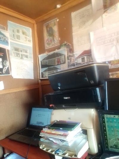 The image shows a room in Auchi, Nigeria (latitude: 7.062, longitude: 6.273). The room is a small office, with corkboard walls covered with architectural drawings and diagrams. In the corner, there are three printers stacked on top of each other. The top printer is black, the middle printer is black and says "Canon" on it, and the bottom printer is white. There are two laptops on the desk, a black one on the left and a black one on the right. The black laptop on the left is open and a document can be seen on the screen. The black laptop on the right is also open and a map can be seen on the screen. There are several books on the desk, stacked on top of the bottom printer. A power cord for the laptop is also on the desk. The room is well-lit. There is no one visible in the image.