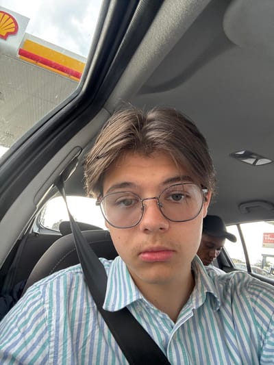 The image shows a young man wearing glasses and a blue and white striped shirt, sitting in the front passenger seat of a car. He is looking directly at the camera. The seatbelt is fastened. The man has short brown hair and is wearing a black belt. In the background, a second person can be seen wearing a black hat, partially obscured. The image appears to have been taken inside a vehicle. The coordinates (latitude: 53.921, longitude: -0.796) place the scene in Pocklington, United Kingdom. The location is likely a petrol station as a Shell sign is visible through the window. The environment is a car interior.
