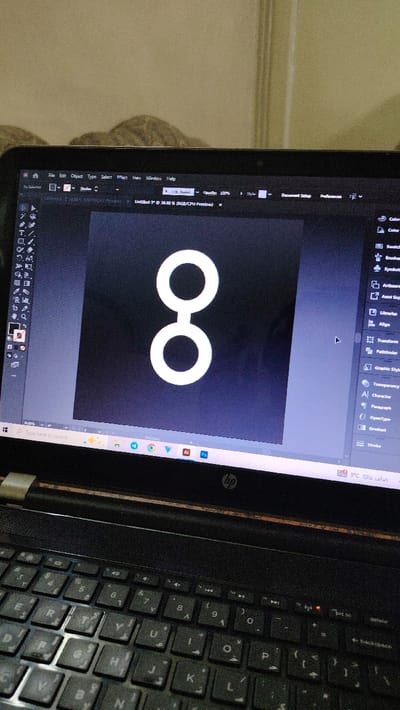 The scene depicts an indoor environment in Al-Qaboun Municipality, Syria, captured from a slightly elevated, oblique perspective looking down at a powered-on black HP laptop.

The laptop's screen is illuminated, displaying the Adobe Illustrator application. The primary workspace within Illustrator features a white graphic resembling a vertically stacked figure-eight or two connected circles, centered against a solid black background. The Illustrator user interface is visible, including top menus (File, Edit, Object, Type, Select, Effect, View, Window, Help), toolbars on the left and right sides of the canvas, and a document tab labeled "Untitled-3* @ 38.86% (RGB/CPU Preview)". A black arrow mouse cursor is positioned on the right side of the Illustrator workspace. The Windows taskbar is visible at the bottom of the screen, displaying several application icons, including Chrome, Edge, Adobe Illustrator, and Adobe Photoshop. The taskbar also shows system information, specifically the Arabic text "صادر عاليًا" (likely related to language settings) and a weather icon indicating "8°C".

The laptop's black keyboard features keys with both Latin and Arabic characters, indicating an Arabic keyboard layout; some keys show visible wear. The HP logo is centered above the keyboard. A small, light-colored rectangular sticker or label is partially visible on the top-left edge of the laptop's body, above the keyboard.

The background consists of a light-colored, textured wall or vertical surface. Directly behind the laptop, a portion of a patterned, possibly fabric, object is visible, consistent with a cushion or furniture backrest. To the upper right, a vertical light-colored strip, possibly part of a door frame or wall corner, is present. No individuals are visible in the scene.