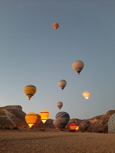 This is a wide-angle, eye-level, outdoor, full shot of several hot air balloons in the sky over a rural landscape, possibly Cappadocia, Turkey, at sunrise. The sky is a clear, light blue. The balloons vary in color and size, with some being brightly lit from within. One balloon in the foreground on the left is yellow with black stripes and the words "Air Kapadokya" written on it. Another balloon to its right is also yellow and lit from within. Further to the right, there are other balloons with various patterns, including a blue and white checkered one on the far right. The ground in the foreground is dry, golden-brown earth, appearing to be a field. In the background, there are rolling hills and rock formations. There are no people visible in the frame.