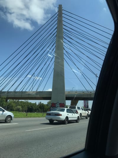 A bridge pylon with attached suspension cables dominates the view, seen from within a vehicle. A second, parallel bridge is visible in the background. Graffiti marks the base of the visible bridge's pylon. A line of white vehicles, including a car directly next to the bridge pillar, are on the road. Green foliage is visible beyond the road. The sky is blue with scattered clouds, as seen through the car's window.
