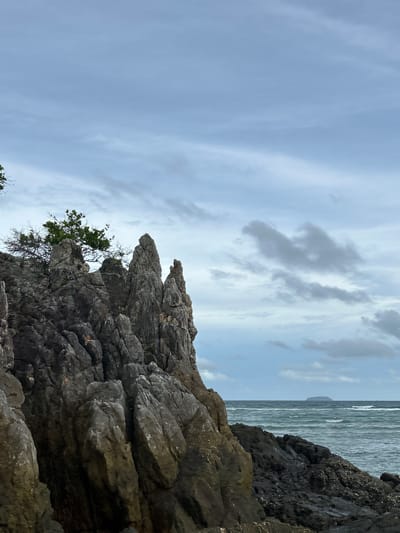This image depicts a rocky coastline with the sea in the background, located in Phru Nai, Thailand. In the foreground, jagged, gray rocks dominate the left side of the frame, with a sparse green tree growing from the top of one of the rock formations. The sea is visible to the right, with choppy, greenish-blue waves. A small, dark island is visible on the horizon. The sky is overcast with a mix of gray and white clouds.