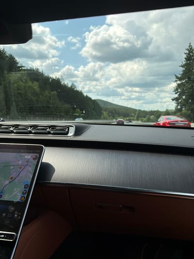 The image, taken from inside a car, shows a view through the windshield. The foreground features the car's dashboard with a navigation screen displaying a map of Studena, Bulgaria. The screen also shows the time as 15:59 and a temperature of 22.0. The dashboard is a dark, textured material. The car is in motion on a highway, as evidenced by the blurred scenery and other vehicles on the road. In the middle distance, several cars are visible, including a red sports car in the right lane. The background consists of a tree-lined hillside and a cloudy sky. The overall impression is a snapshot of a car journey through a scenic route near Studena, Bulgaria.