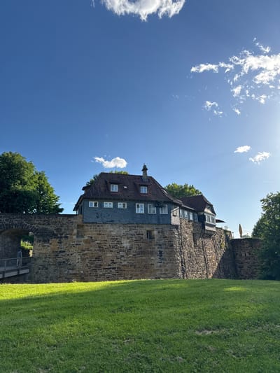The image depicts a view of the Esslingen Castle (Esslinger Burg) in Esslingen am Neckar, Germany. The castle is situated atop a hill and features a prominent stone wall. A building with a dark, shingled roof and multiple windows is visible behind the wall. The foreground consists of a well-maintained green lawn, with a path and archway leading into the castle grounds on the left side of the image. A person is walking across the path. The sky is clear and blue with scattered white clouds, and the sun is shining brightly, casting shadows on the lawn and wall. Lush green trees surround the castle structure.