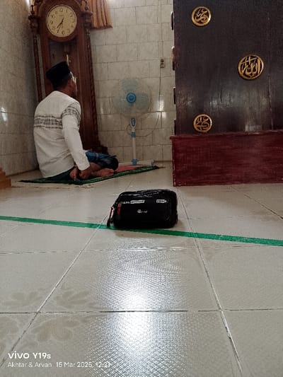 The image shows a man kneeling on a prayer rug in a room with white tiled walls. He is wearing a white shirt and black pants. He is facing away from the camera, with his head bent in prayer. On the floor next to him is a black bag and a green tape line. In the background, there is a wooden cabinet with gold Arabic script on it. A standing fan is also visible, as well as an antique clock on the wall to the left of the image. The room appears to be a mosque or prayer room in Kecamatan Anjatan, Indonesia, based on the provided coordinates. The man appears to be engaged in prayer, with the environment reflecting a religious context. The black bag and green tape suggest there may be a gathering or activity happening within the mosque.