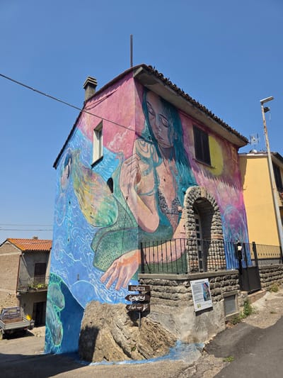 The image depicts a mural on the side of a building in Viterbo, Italy, featuring a mermaid with flowing teal hair and a scaly green tail. The mermaid is depicted with her eyes closed, holding her hands to her chest, and is surrounded by vibrant colors of pink, yellow, and blue, with wavy patterns representing water. An arched doorway made of stone is visible on the building, with a black metal fence in front of it. A signpost with directional arrows points towards "Sirenetta" and "Gemerotola." The building's facade is painted with various other artistic elements, including what appears to be a sun or moon on the right side of the mural. In the background, other buildings, a street lamp, and a clear blue sky are visible. The mural is a prominent feature of the street scene, dominating the side of the building.