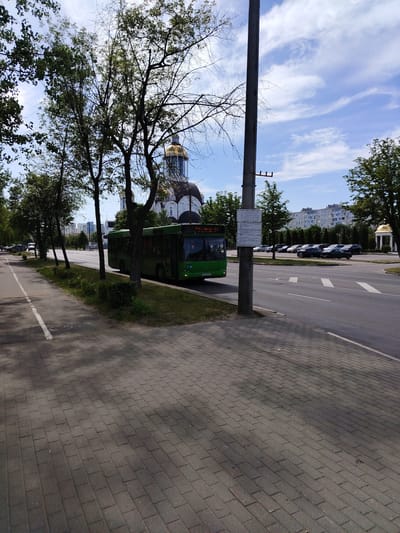 The image depicts a street scene in Salihorsk, Belarus, on a bright, partly cloudy day. A green public bus is driving on a multi-lane road, with a sidewalk made of paving stones on the left. Trees line the sidewalk and the median of the road, with lush green leaves suggesting it is late spring or summer. In the background, a large building with a golden dome, likely a church or cathedral, is visible through the trees. Further in the distance, apartment buildings can be seen. A tall, grey utility pole stands prominently on the right side of the image, with a sign attached to it. Cars are parked on the side of the road and a few are in motion. The overall atmosphere is that of a typical urban environment on a pleasant day.