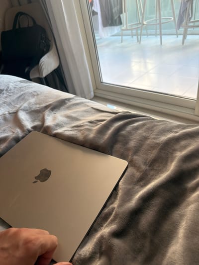 The image depicts an indoor scene within Business Bay, United Arab Emirates, observed from a low perspective. A closed silver Apple MacBook with its iconic logo is positioned on a rumpled, textured grey blanket or duvet, which dominates the foreground and midground. A person's fair-skinned hand is partially visible in the bottom-left corner, resting near the laptop. In the background, a large glass window or sliding door provides a view of an outdoor tiled balcony. Several white high stools are visible on the bright white tiled floor of the balcony. To the left of the window, a light-colored chair holds a black bag with a visible strap. White curtains are partially drawn along the left side of the window frame. Natural light enters from the window, illuminating the scene and creating variations in shadow and highlight on the blanket's texture.
