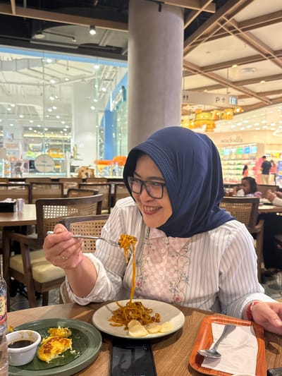 A woman wearing a navy blue hijab and glasses is eating noodles at a restaurant in Gunung Putri, Indonesia. She is holding a fork with noodles and is looking at her food. There are plates of food on the table, including noodles, rice cakes, and crackers. The restaurant appears to be brightly lit and decorated with wooden accents. In the background, there are other diners and store fronts visible through a large window. A dog is visible in the background through the window, appearing to be a golden retriever.