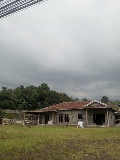 A house under construction stands on a grassy lot under a cloudy sky. The house has a red-tiled roof and cement walls with window openings. Trees line the background, adding a touch of nature to the scene. Power lines are visible in the upper left corner. The atmosphere is overcast and somewhat dreary.
