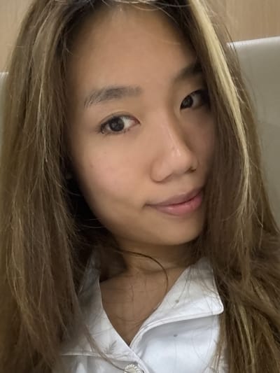 A close-up portrait captures the face of a young person, likely female, with long, wavy, light brown hair featuring distinct blonde highlights. Her face, oriented sideways, looks directly into the camera with wide, dark eyes and a calm, subtly smiling expression, her lips slightly parted. Strands of her hair fall across her forehead and frame her face, some partially obscuring her left eye. She is wearing a crisp white collared shirt, possibly a uniform, with a decorative button visible near her right shoulder. The background is softly blurred and indistinct, suggesting an indoor setting. The image conveys a relaxed and direct gaze. No text is visible in the image. The photo's location context is Dunhuang City, China.