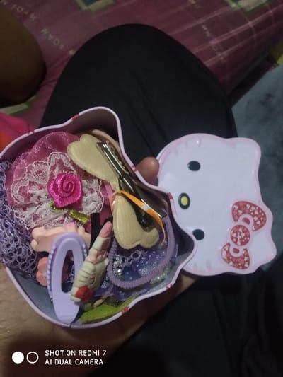 A partially-open, light pink Hello Kitty container overflows with hair accessories. Lace, rose-adorned fabric, various barrettes, hair clips, and elastic bands are visible. The lid, also Hello Kitty themed, is slightly ajar, exposing the container's contents. The scene is set against a background of clothing or bedding in shades of black, pink and gray.