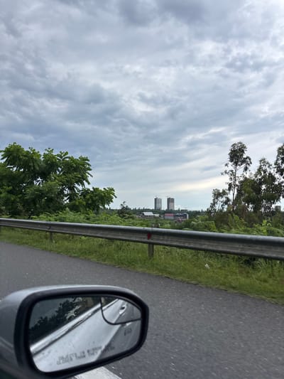 This image depicts a view from a vehicle traveling along a road in Nakaidzeebi, Georgia. The foreground shows the asphalt road and the driver's side mirror, reflecting trees and sky. A metal guardrail runs along the side of the road, separating it from a grassy embankment and lush green vegetation. In the middle ground, a cluster of buildings and trees is visible, with two tall, unfinished high-rise buildings dominating the horizon. The sky is overcast with gray clouds.