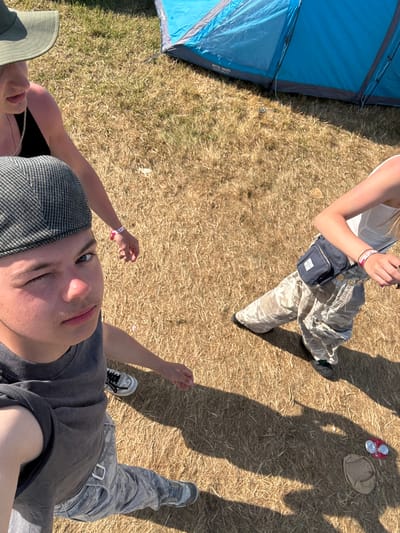 An overhead shot shows three people on dry grass next to a blue tent. One person wears a white top and camo pants with a small blue bag. Another wears a green hat and a black top. The third person wears a grey hat and shirt with camo pants. Their shadows are visible on the grass next to two empty beer cans and a hat.