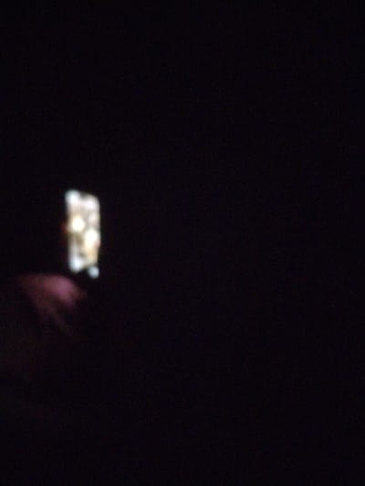 The image depicts a very dark scene with a single, out-of-focus rectangular object emitting light on the left side. The object appears to be a window or a screen with some light sources visible within it. Below and to the left of this lit object, there is a blurred, pinkish-purple mass, which could be a hand or part of a person's clothing, partially obscuring the lower part of the lit object. The background is predominantly black, suggesting a nighttime setting or a very dimly lit interior. There is no discernible information in the image that would allow for the identification of its specific location within the city of Ștefan Vodă, Moldova. The scene is devoid of any clear details about people, specific actions, or interactions.