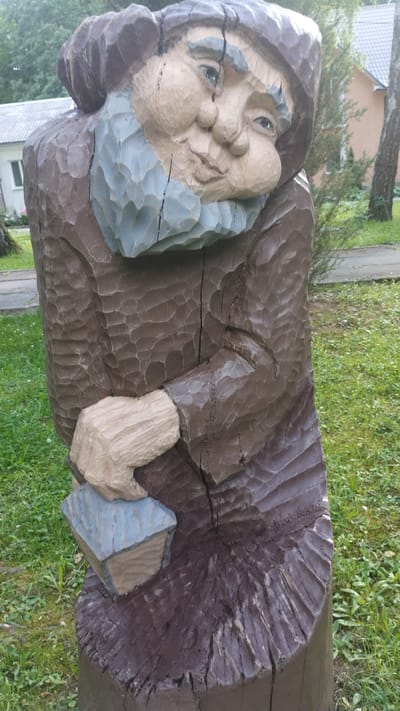 The image shows a wooden sculpture of a gnome or an old man carved from a tree trunk. The sculpture is painted in brown and blue. The figure has a large, round face with chubby cheeks, a prominent nose, and a wide smile. It is wearing a hooded robe with a blue scarf around its neck. The sculpture is holding a small, rectangular object, possibly a book or a box, in its left hand. The sculpture is located outdoors in a grassy area with some trees and buildings in the background, suggesting a park or public space. The location is identified as Khmilnyk, Ukraine.