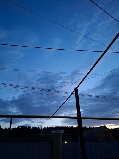 The image depicts a view of the sky with power lines and a utility pole in the foreground, possibly located in the city of Амзя, Russia. The sky is a gradient of blue hues, with some clouds visible. The power lines are dark and extend across the frame. A utility pole is present on the right side of the image, appearing metallic and somewhat obscured by shadows. In the background, there's a suggestion of trees and a building or structure, all under dark conditions that suggest evening or twilight.