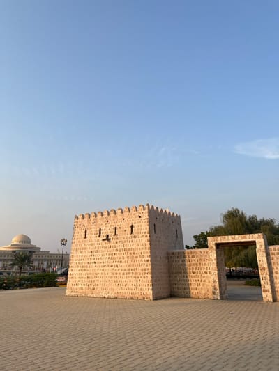 The image depicts a historical stone fort structure in Sharjah, United Arab Emirates, specifically located within the Sharjah Heritage Area, near Bank Street. The central feature is a light-colored, square-plan, stone-built fort with a crenellated parapet roofline and several small, rectangular openings (embrasures) on its visible faces. Attached to the fort's right side is a lower stone wall segment, which leads to a freestanding stone archway providing access to an area beyond. The foreground consists of a large, light-grey paved plaza, constructed with interlocking pavers. In the distant left background, a modern building with a prominent domed roof and multiple stories is visible, along with green landscaping and palm trees. To the right of the fort and through the archway, dense green foliage from trees is present. The sky above is clear and light blue with minimal, wispy clouds. No individuals or vehicles are present within the frame. The scene is illuminated by natural light, casting soft shadows.