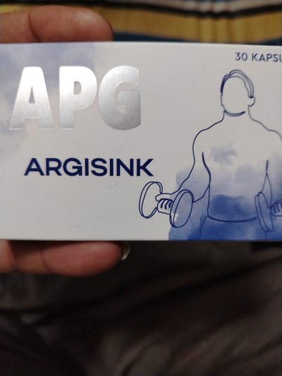 A close-up of a product box for "APG ARGISINK" is pictured. The box has a white background and features the large, metallic silver letters "APG" at the top left. Below this, in dark blue text, is "ARGISINK". To the right of the text, a minimalist blue line drawing depicts a muscular male torso holding dumbbells, suggesting a fitness or supplement product. The top right corner of the box shows the text "30 KAPSUL". The box is held by a person's fingers in the foreground, partially obscuring the bottom of the image. The background consists of out-of-focus fabric in muted yellow and brown tones. There is no indication of the city of Khiva, Uzbekistan, in the image.