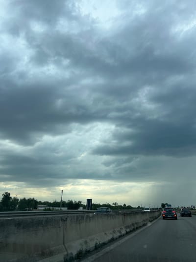 The image depicts a view from a vehicle traveling on a highway, likely the SS16bis, near Bisceglie, Italy. The sky is overcast with dark, heavy clouds, suggesting impending rain, with a visible streak of rain falling in the distance on the right side of the frame. The highway has a concrete divider separating traffic, with several cars visible on the road ahead. Trees and some buildings are present in the background beyond the divider. The road surface is asphalt.