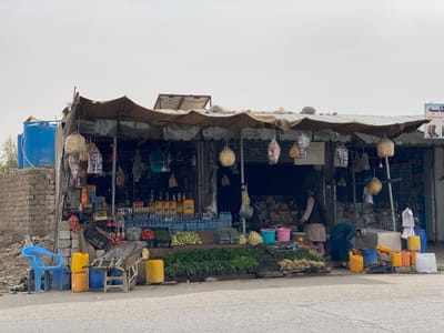 The scene is a small, open-air shop, located in the Panjwai district of Kandahar, Afghanistan, at the coordinates 31.542° N, 65.463° E.  The shop is constructed of corrugated metal and wood, with a corrugated metal roof. It is shaded by a brown canvas awning supported by wooden poles. There are various items hanging from the awning, including woven straw balls, plastic bags, and what appear to be food items.  The shop has a display of merchandise for sale, including bottles of water, food products, and fresh produce, including leafy green vegetables and tomatoes. Two women are standing in front of the shop, one in a blue burqa and the other in a long blue dress. A third woman is crouched down by a blue cooler on the right side of the shop. Behind the shop, there is a wall of concrete blocks and a blue water tank.  There are also various objects in front of the shop, including yellow buckets, blue chairs, and baskets of produce.  The ground is dusty and uneven, and the sky is overcast.
