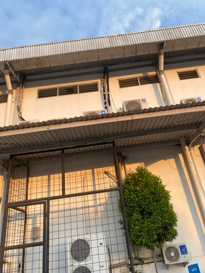 A low-angle exterior shot captures the side of a modern industrial building in Gambut Barat, Indonesia, bathed in the warm glow of late afternoon sunlight. The building features a corrugated metal roof extending over a section of the wall, beneath which several air conditioning units are mounted. Above this overhang, a row of horizontal slatted windows is visible on the white facade. Dark pipes and electrical conduits run vertically along the building's exterior. In the foreground, a metal security cage with a gate partially obscures the lower portion of the building and a large, leafy green bush. The sky above is a clear blue with wispy clouds.