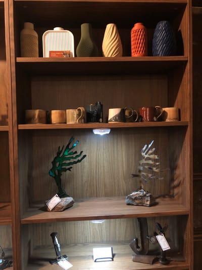 The image shows a wooden bookshelf displaying various decorative items. On the top shelf, from left to right, are a beige vase with a geometric pattern, a white ribbed rectangular object with a business card on it, a green ribbed vase, a cream-colored ribbed vase, an orange ribbed vase, and a dark blue ribbed vase. The business card on the white object reads "Susan Walker-Ing."

The second shelf from the top contains four ceramic mugs. The left two appear to be a gradient of brown and beige. The third mug from the left is a golden beige, and the rightmost mug is dark blue with a glossy finish.

The third shelf from the top features two metallic sculptures. On the left is a green, branched sculpture resembling coral or an abstract tree. On the right is a metallic sculpture of a tree with a lighter, more detailed branch structure. Both sculptures are placed on rough stones.

The bottom shelf displays three metal objects. On the left is a dark metal object that appears to be part of a larger structure or tool. In the center is a sculpture made of rusted metal pieces, possibly abstract, with a white tag attached. On the right, a similar rusted metal sculpture is visible with a white tag.

The environment appears to be an indoor display area with wooden shelving units. The lighting is soft, with some visible light sources illuminating the objects on the second and third shelves. The overall aesthetic suggests a retail space or a curated collection of art and decor. The city of Severn, Canada, is not explicitly mentioned in the image or its contents.