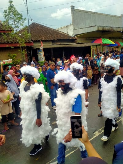 The image captures a public procession or cultural event occurring on a street in Mojogedang, Indonesia, under an overcast sky. Four individuals are prominently featured in the foreground, advancing from left to right. Each is clad in an identical white, voluminous, shaggy costume, resembling a sheep or a cloud, paired with a large, white, fluffy headpiece. Their faces are entirely covered with dark, possibly black, paint or masks. Underneath the costumes, one figure wears a black long-sleeved shirt, another a blue short-sleeved shirt, and two wear black short-sleeved shirts. In the bottom right corner, a human hand holds a black smartphone, oriented vertically, pointed towards the costumed figures, indicating recording or photography. A dense crowd of spectators, encompassing both adults and children in various casual attire, lines both sides of the thoroughfare, their gazes directed towards the procession. The background features architectural elements: a building with a traditional tiled roof on the left, partially obscured by foliage, and a multi-story concrete structure on the right. Beneath the concrete building, market stalls are visible, characterized by vibrant multi-colored umbrellas (red, yellow, green, blue) and displayed merchandise. The street surface is dark, appearing to be paved.