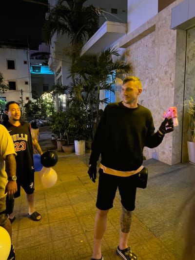 The image shows an outdoor scene in an urban setting. A blonde man stands in the top of the image wearing black gloves, a black long-sleeved shirt, and black shorts, holding what appears to be a pink toy gun. A man wearing a Chicago Bulls jersey stands in the lower part of the image holding a cluster of balloons. The balloons are blue, black, and white. In the background, a building with trees is visible, indicating the location as a street in Unknown, Vietnam. The street is paved with textured bricks.