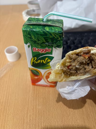 The scene depicts a close-up, elevated view of food and beverage items on a light-colored wooden surface, located in Adrogué, Argentina. In the center-left, a "Baggio Pronto Naranja" orange juice box, predominantly green with leaf patterns and orange fruit graphics, stands upright. A green and white striped plastic straw is inserted into the top of the juice box, with its flexible segment bent. To the right, a partially consumed empanada, held by an unidentified hand, is presented. The empanada's bite-revealed interior shows a filling of ground meat, visible red pepper pieces, and lighter-colored elements possibly indicative of potato or egg. It is partially wrapped in a crumpled white paper napkin. In the blurred background, a white cylindrical object is visible to the left of the juice box, and a black computer keyboard is present to the right. Further back, out-of-focus white and grey objects are distinguishable.