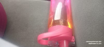 A translucent plastic water bottle with a pink-to-orange gradient occupies the right and center portions of the frame, resting on a light gray, textured fabric surface. The bottle features a pink cap assembly, which includes a textured cylindrical grip and a curved carrying handle. Faint measurement markings, including "32OZ," are visible vertically on the bottle's side. A blurred, dark pink or red cylindrical object is partially visible on the far left. The scene is illuminated, creating reflections on the bottle. No individuals or discernible actions are present. The bottom right corner of the image contains a digital watermark: "Samsung Michael's A05 September 23, 2025." There are no visual indicators within the image to confirm the location as Uweyoba, Nigeria.