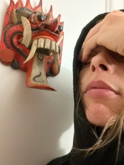 The image displays a rotated view of a person and an ornate mask against a plain, light-colored background. In the upper portion of the frame, a person's face is visible, oriented upside down. Features include their nose, mouth with slightly parted lips, and part of their chin. Strands of blonde hair are visible, and the person is wearing a dark hoodie. One hand covers their forehead and eye, partially obscuring their face.
Below the person, also inverted relative to a standard upright view, is a red, traditional-style mask or carving. This mask is highly decorative, featuring prominent white fangs or teeth, black circular eyes, and intricate patterns, including black and white swirls, applied over a vibrant red base. The design suggests it might be a cultural or ceremonial piece.
The scene appears to be set indoors, likely under artificial lighting. The unconventional, inverted perspective suggests the photo was taken from an unusual angle, possibly a selfie or an overhead shot. There is no visible text in the image. While the location context is Vicente López, Argentina, the visual details within the image do not provide specific cues to confirm this. The person's expression is partially concealed by their hand, creating an ambiguous or contemplative mood.