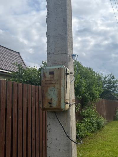 A weathered, rusted metal electrical box is attached to a concrete utility pole. The box has Cyrillic text on it which reads "702", "НАПРЯЖЕНИЕ ПОД", and "НЕ ОТКРЫВАТЬ". A black electrical cable descends from the box and is secured to the pole. A small blue and white object with wires is attached higher up on the pole. In the background, a brown wooden fence runs along the side of the property, with lush green trees and bushes behind it. A portion of a house with a brown shingled roof is visible to the left. The ground is covered in green grass. The sky is overcast with white and gray clouds. The scene appears to be in a rural or suburban setting, possibly in the village of Borodulino, Russia, given the Cyrillic text. There are no people visible in the image.